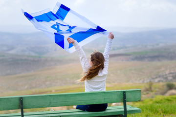 Beautiful young jewish girl sitting holding Israel flag in the wind and enjoying great view landscape on the sky, field and mountains.Patriotic holiday. Independence day Israel - Yom Ha'atzmaut .