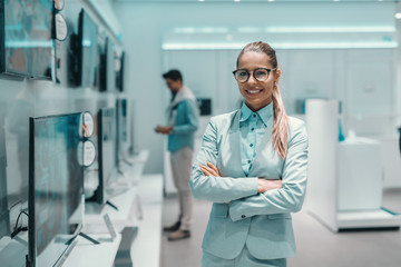 Fototapeta premium Smiling Caucasian brunette in formal wear and with eyeglasses standing next to tv with arms crossed in tech store.