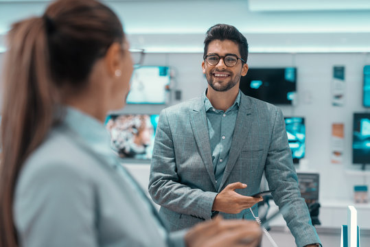 happy mixed race unshaven man in formal wear trying out new smart phone and looking at his lovely wife. Tech store interior.