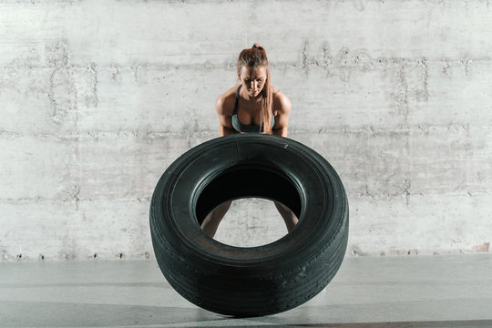 Strong dedicated female bodybuilder flipping tyre in crossfit gym. In background wall. Full length.