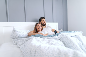 Obraz na płótnie Canvas Young smiling happy attractive Caucasian couple lying in bed and watching television in the morning. Bedroom interior.