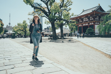 full length asian tourist woman with backpack at vacation walking through shitennoji japanese temple in osaka japan. girl travel with professional dslr camera relaxing enjoy sunshine outdoors.