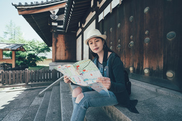 Young pretty girl in straw hat with tourist map sitting on the steps of shitennoji temple. beautiful female traveler holding paper tour guided book reading consider finding direction rest on stairs.