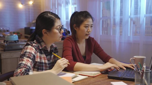 two girl roommates team workers having discussion while doing report on final exam using laptop computer at home kitchen in midnight. friends talking pointing screen pencil another typing on keyboard