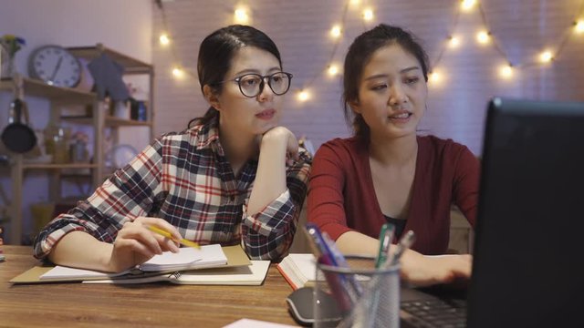 Group of university girl students working together on final project semester. young college female using laptop computer having discussion talking at night indoors. two hard-working woman at home.