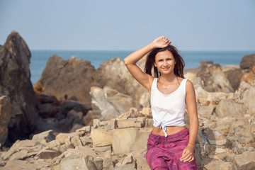 A smiling young woman with hair fluttering in the sea wind holds her hand over her head and squints at the bright sun against the sea and rocks