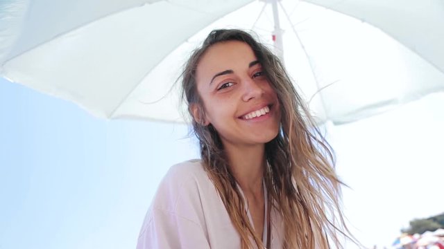 beautiful tanned happy laughing woman is sitting on the beach. woman in white t-shirt enjoing and lying under big white beach umbrella on the sunny beach at summer vacation. HD Stock video footage