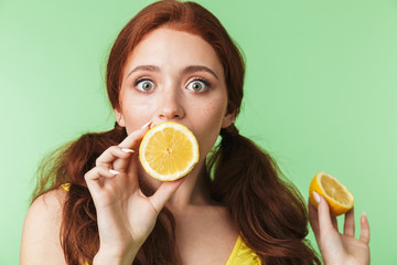 Shocked young redhead girl posing isolated over green wall background with citrus vitamins fruits.