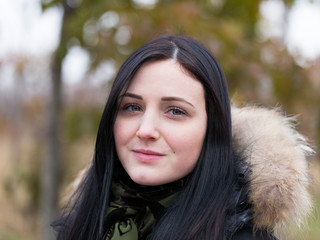 Horizontal closeup of pretty young woman with long straight dark hair and porcelain skin in fur-trimmed coat standing in front of soft focus Fall foliage