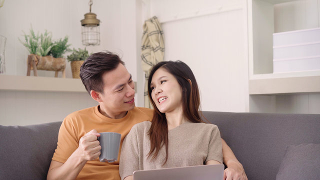 Asian couple using laptop and drinking warm cup of coffee in living room at home, sweet couple enjoy love moment while lying on the sofa when relax at home. Lifestyle couple relax at home concept.