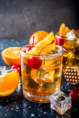 Classic alcohol cocktail, whiskey with ice cubes, orange slices and cocktail red cherries. In two glasses, on a dark blue concrete table,