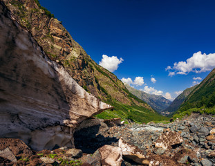 Fototapeta premium A huge block of snow melts in the summer in the Caucasian mountains near Dombai and a stream flows nearby.
