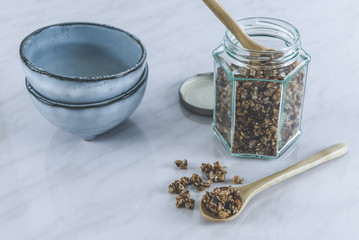 Homemade healthy and nutritious breakfast granola in a glass jar with wood spoon and blue ceramic bowls on clean marble kitchen table in fresh morning light