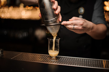 Professional bartender pouring cocktail from the steel shaker through the sieve