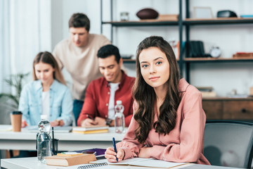 Pretty student writing in notebook and looking at camera