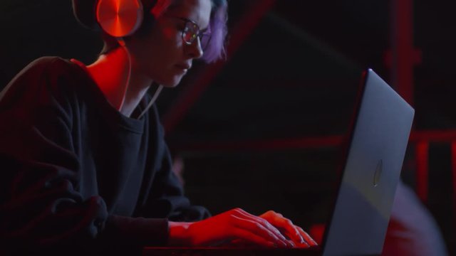 Medium shot of teenage girl sitting at table in headphones, listening to music and typing on laptop keyboard