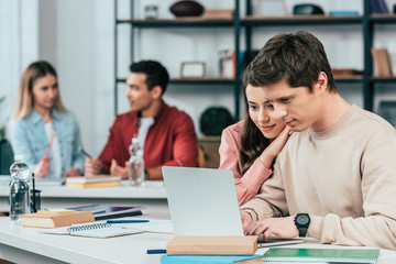 Smiling students sitting at desks and using laptop while studying in classroom