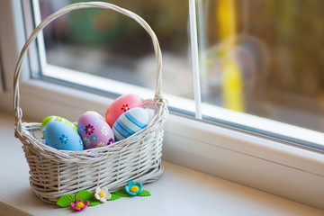 Variety of decorated colorful Easter eggs in wicker basket with spring flowers near window in daylight.