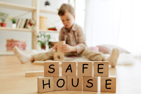 Wooden cubes with words SAFE HOUSE in hands of happy smiling little boy at home. Conceptual image about child rights, education, childhood and social problems.