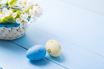 Two painted blue and yellow Easter eggs near basket with white spring flowering branch on light blue background