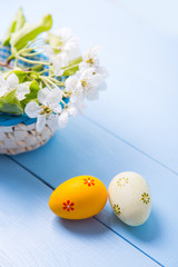 Two painted orange and yellow Easter eggs near basket with white spring flowering branch on light blue background