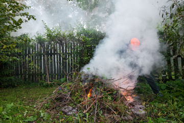 Man in work clothes is working in garden, burning old grass, leaves,  branches after tree pruning and feeding outdoor bonfire in spring or autumn.