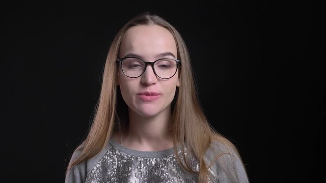 Closeup shoot of young attractive hipster female in glasses showing a tongue and playing a fool looking straight at camera with background isolated on black