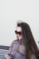 Naklejka premium beautiful girl with dark hair and sunglasses sits on a bench and writes sms in city street