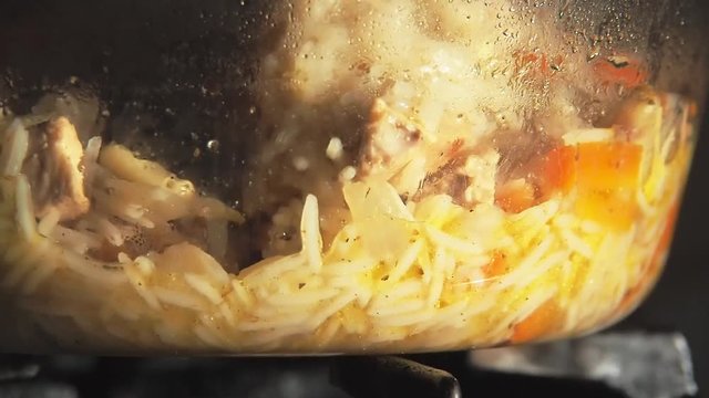 Cooking of food, rice porridge with vegetables and meat. Stirring porridge with wooden spatula. Boiling pilaf in glass pot. Close-up view. Rice with carrots, garlic, onion and meat