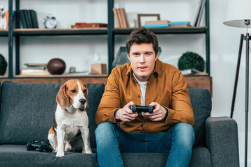 Young man with dog sitting on sofa and holding joystick