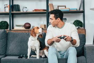 Smiling young man with gamepad sitting on sofa and looking at dog - Powered by Adobe