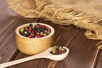 Lot of whole peppercorns of four colors with wooden bowl on jute cloth with wooden spoon on brown wood