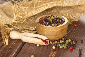Lot of whole peppercorns of four colors with wooden bowl on jute cloth with wooden scoop on brown wood