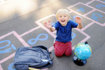 Cute blond boy playing hopscotch game after school with bags laying near