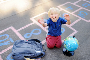 Naklejka premium Cute blond boy playing hopscotch game after school with bags laying near