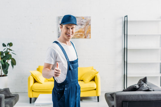 smiling mover in uniform looking at camera and showing thumb up sign in apartment