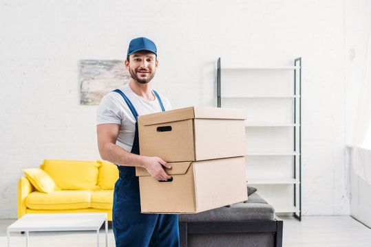 handsome mover in uniform looking at camera while carrying cardboard boxes in apartment with copy space