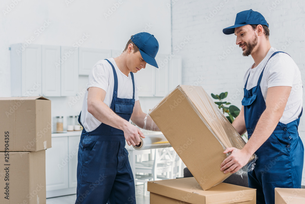 Wall mural handsome movers wrapping cardboard box with stretch film in apartment