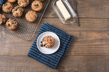 Homemade Blueberry Muffins on Wooden Table Top; One Isolated on White Plate on Blue Kitchen Towel; More Muffins on Cooling Rack in Background; Butter Dish and Knife beside the Muffin 