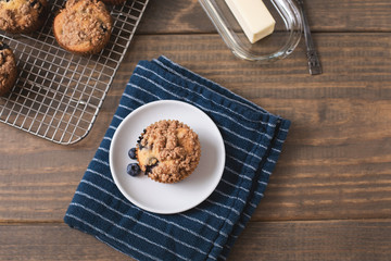 Homemade Blueberry Muffins on Wooden Table Top; One Isolated on White Plate on Blue Kitchen Towel; More Muffins on Cooling Rack in Background; Butter Dish and Knife beside the Muffin 