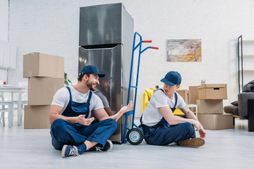 two movers talking while sitting near hand truck, carboard boxes and refrigerator in apartment