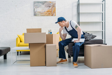 pensive mover sitting near cardboard boxes and holding clipboard in apartment