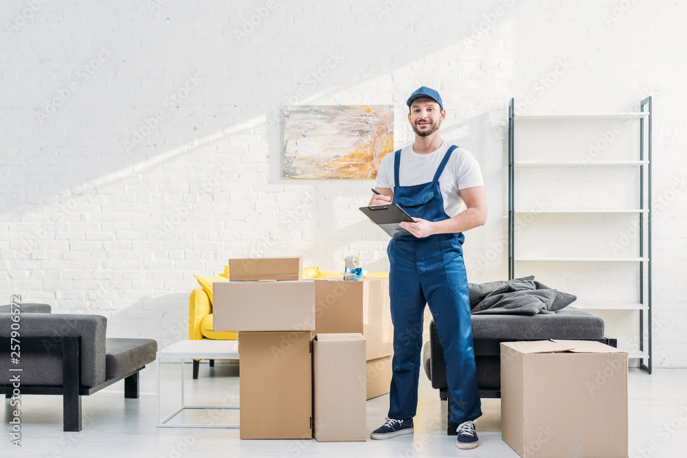 Wall mural mover looking at camera and holding clipboard near cardboard boxes in apartment with copy space