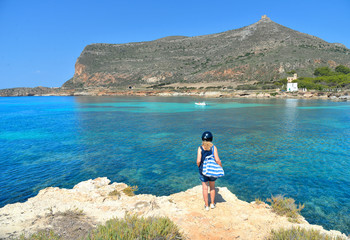 Fototapeta na wymiar woman on the top of the rock cliff looks at paradise clear torquoise blue water with boats and cloudy blue sky in background in Favignana island, Cala Rossa Beach, Sicily South Italy.