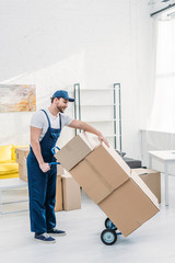 mover in uniform transporting cardboard boxes on hand truck in apartment with copy space