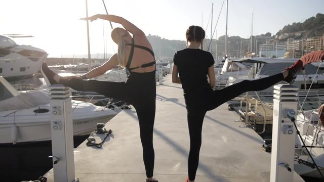 Young women doing exercises at sea port. Fit attractive caucasian girls warm up doing stretching on the pier