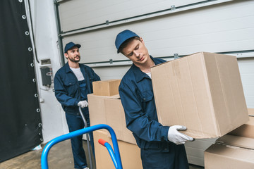 two concentrated movers in uniform transporting cardboard boxes in warehouse