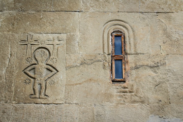 The wall of the church with a small window. An image of an alien or person with crosses, embossed in stone. Georgia, Mtskheta. Religion, Christianity, Orthodoxy.