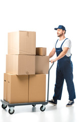 handsome mover in uniform transporting cardboard boxes on hand truck isolated on white