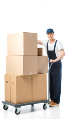 handsome mover in uniform smiling while transporting cardboard boxes on hand truck isolated on white
